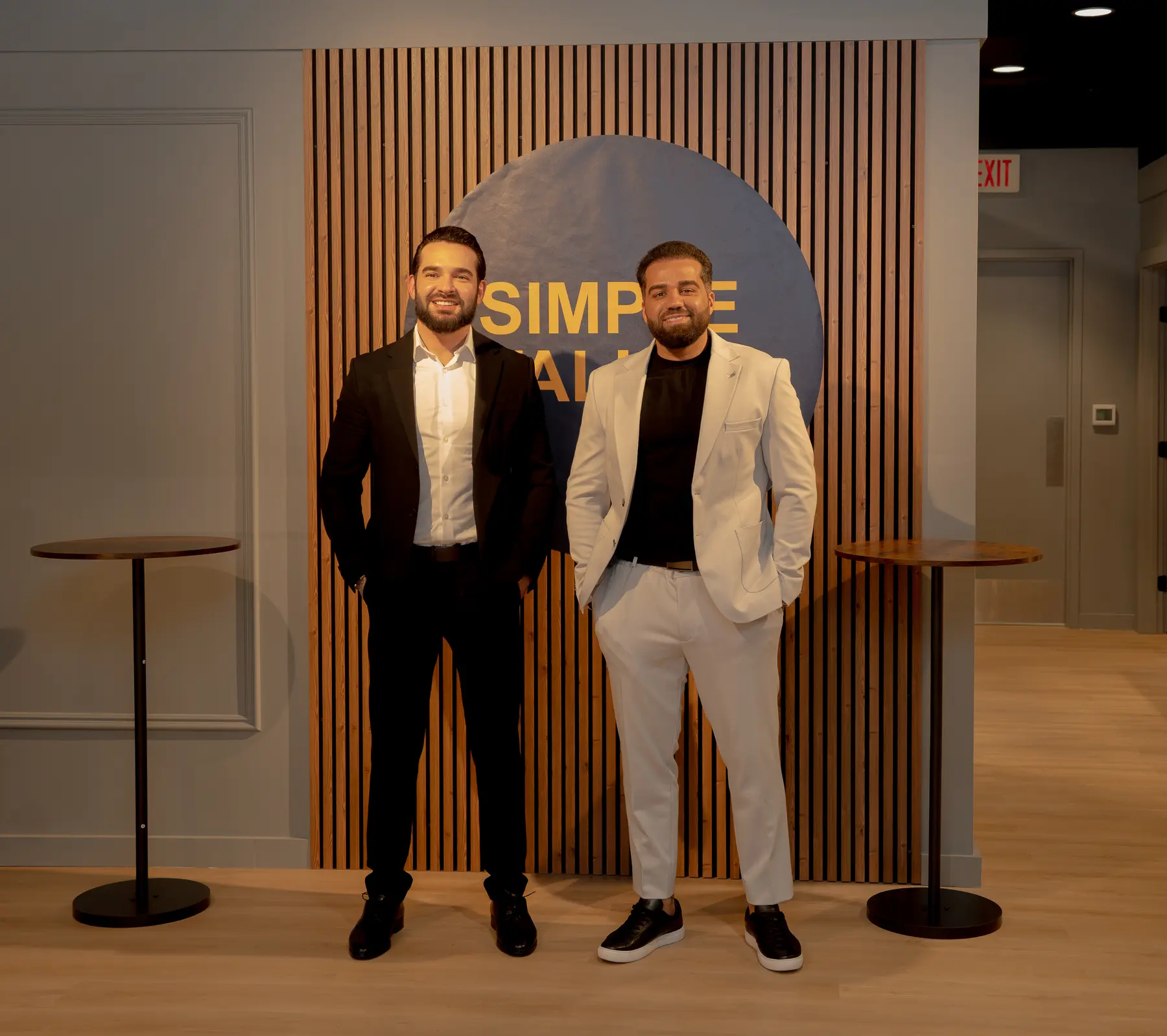 Rafael Pontes e Felipe Drumond em pé diante do painel com o logo do Simple Talk, no estúdio